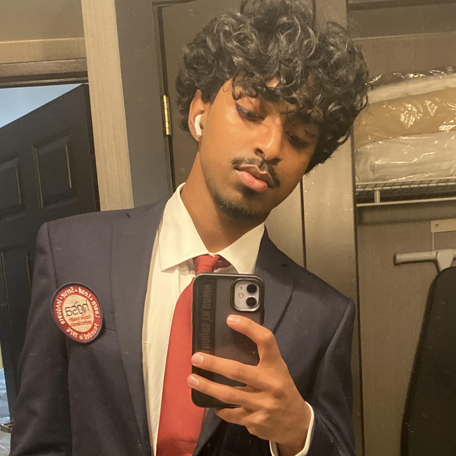 Aadi taking a mirror selfie in a navy suit with a red tie, wearing an earbud and holding a phone in a bathroom.