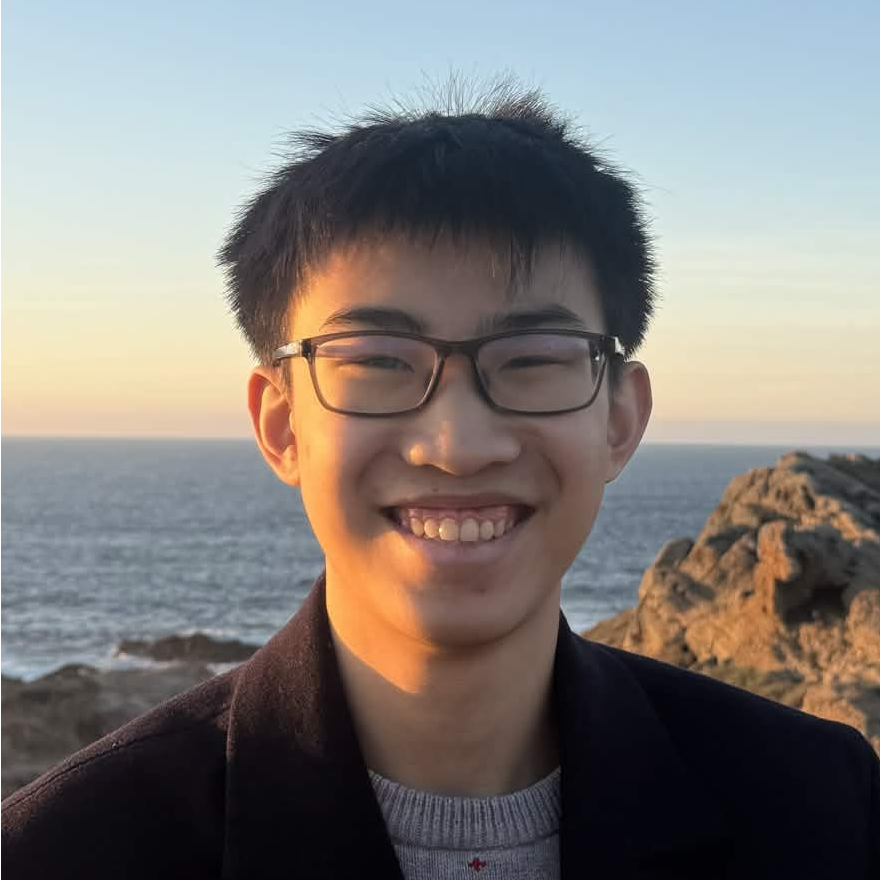 Aaron smiling and wearing glasses in a close-up portrait, with the ocean and rocky coastline in the background at sunset.
