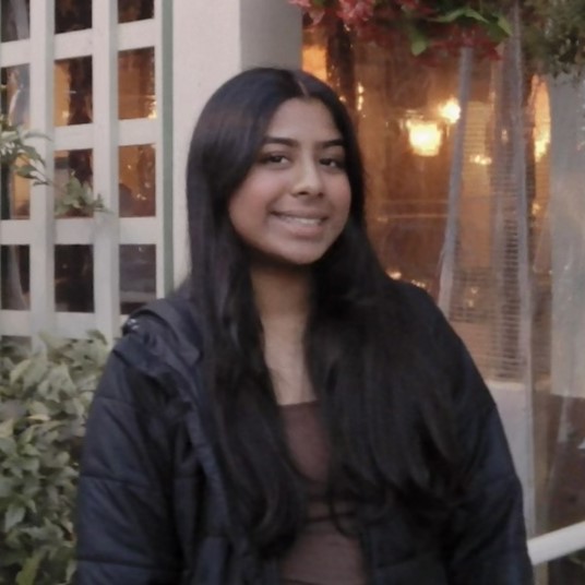 Adithi smiling with long dark hair wearing a black jacket, standing outdoors near a window with warm lights and plants.