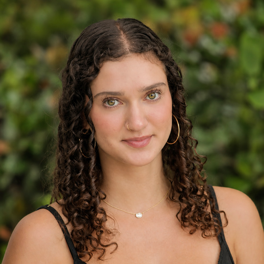 Close-up outdoor portrait of Alexis with curly dark hair, wearing gold hoop earrings and a small nose ring, against a blurred green background.