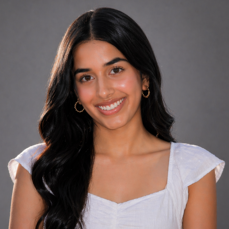 Aneet smiling with long dark hair wearing gold hoop earrings and a white top against a gray background.
