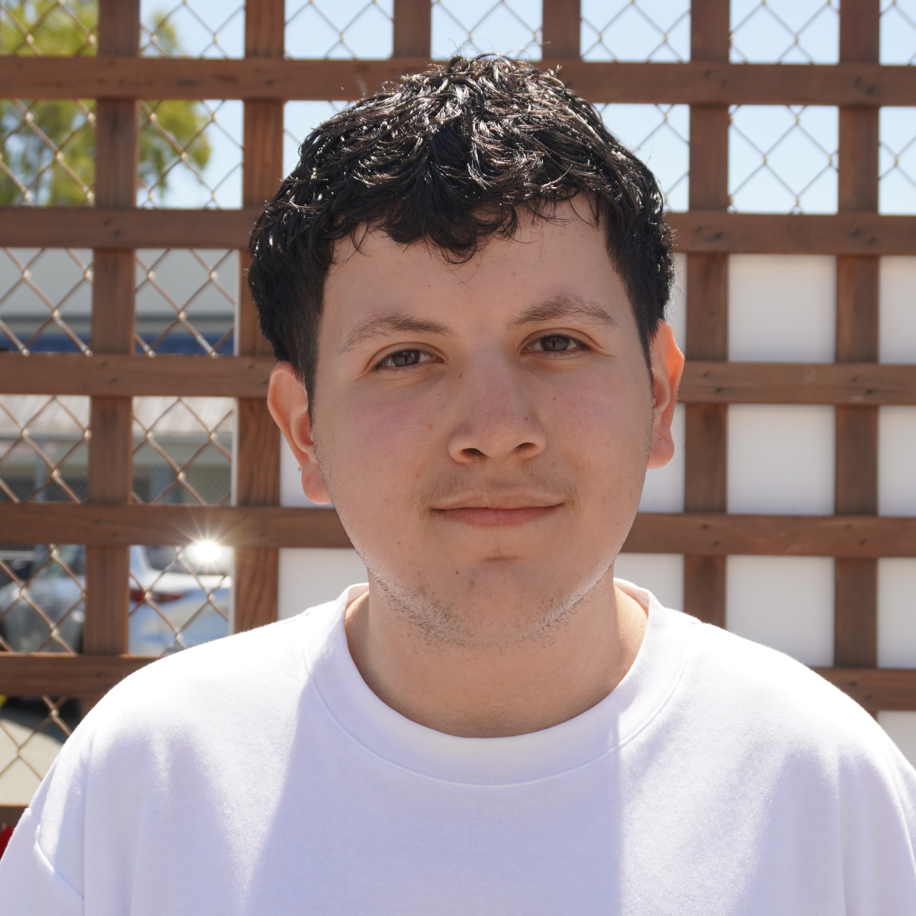 Close-up portrait of Angel with curly dark hair wearing a white shirt, standing in front of a wooden lattice fence in bright sunlight.