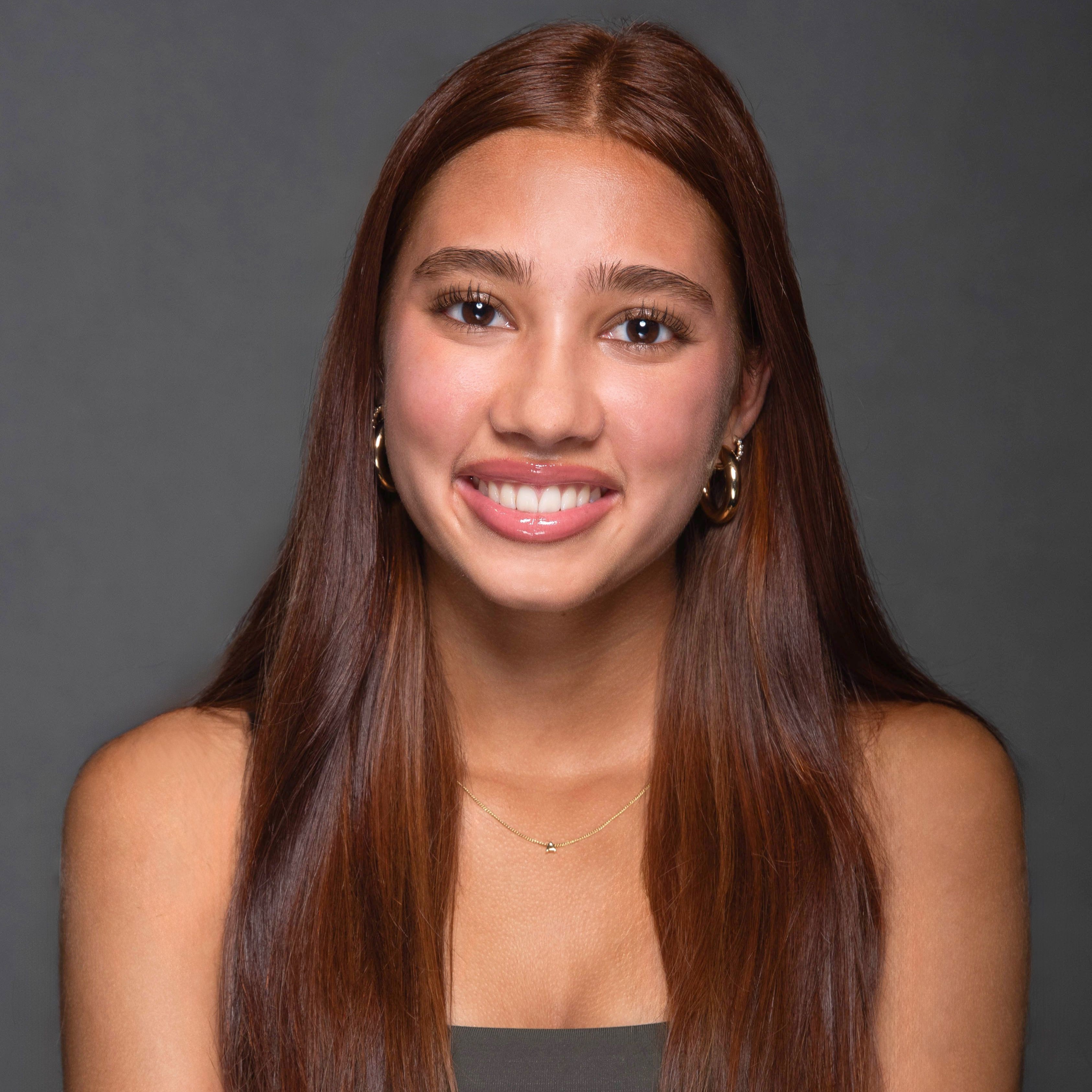 Close-up studio portrait of Anika with long auburn hair, wearing gold hoop earrings and a delicate necklace against a gray background.