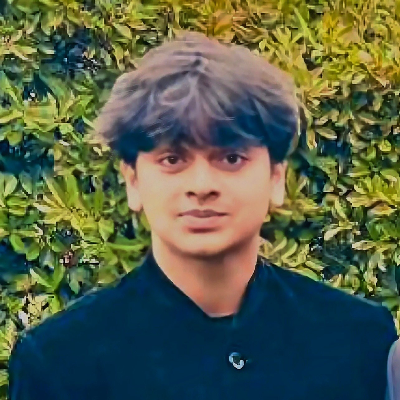 Portrait of Arhaan with dark, wavy hair wearing a dark buttoned jacket, standing in front of leafy green plants.