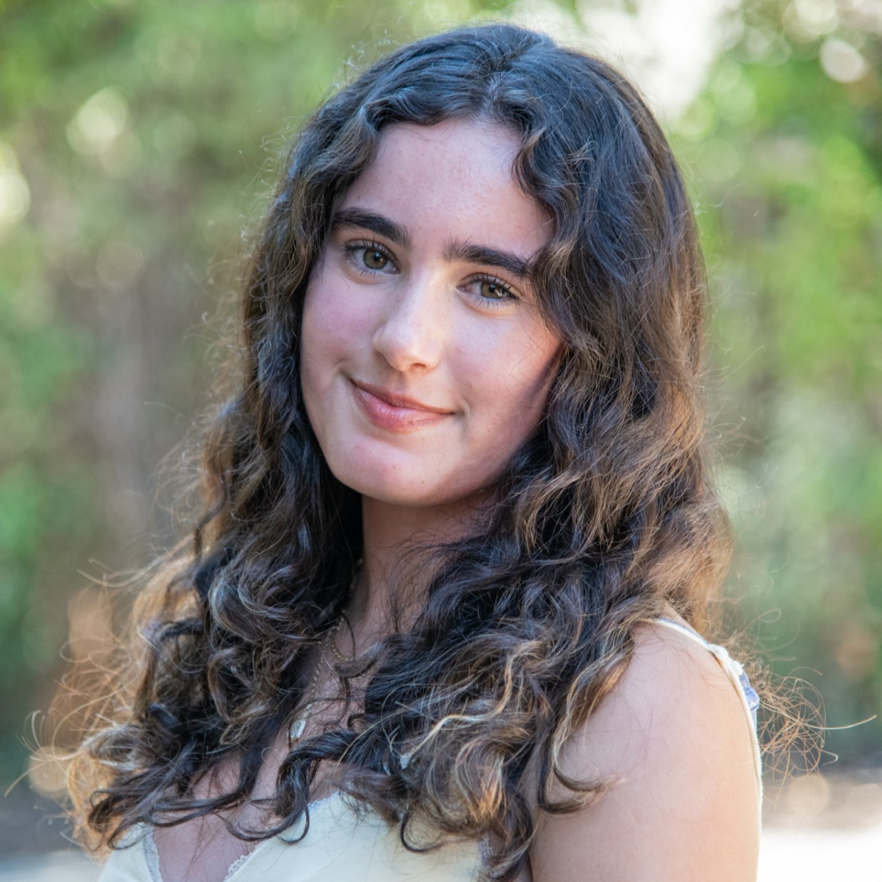 Close-up outdoor portrait of Avery with long curly brown hair, smiling softly against a blurred green background.