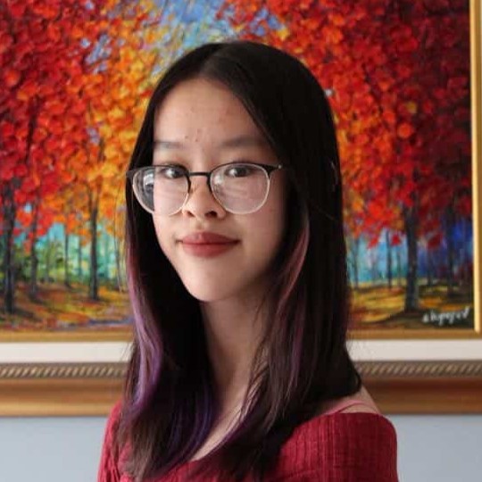 Portrait of Cassie with long dark hair and glasses, standing in front of a framed colorful autumn landscape painting.