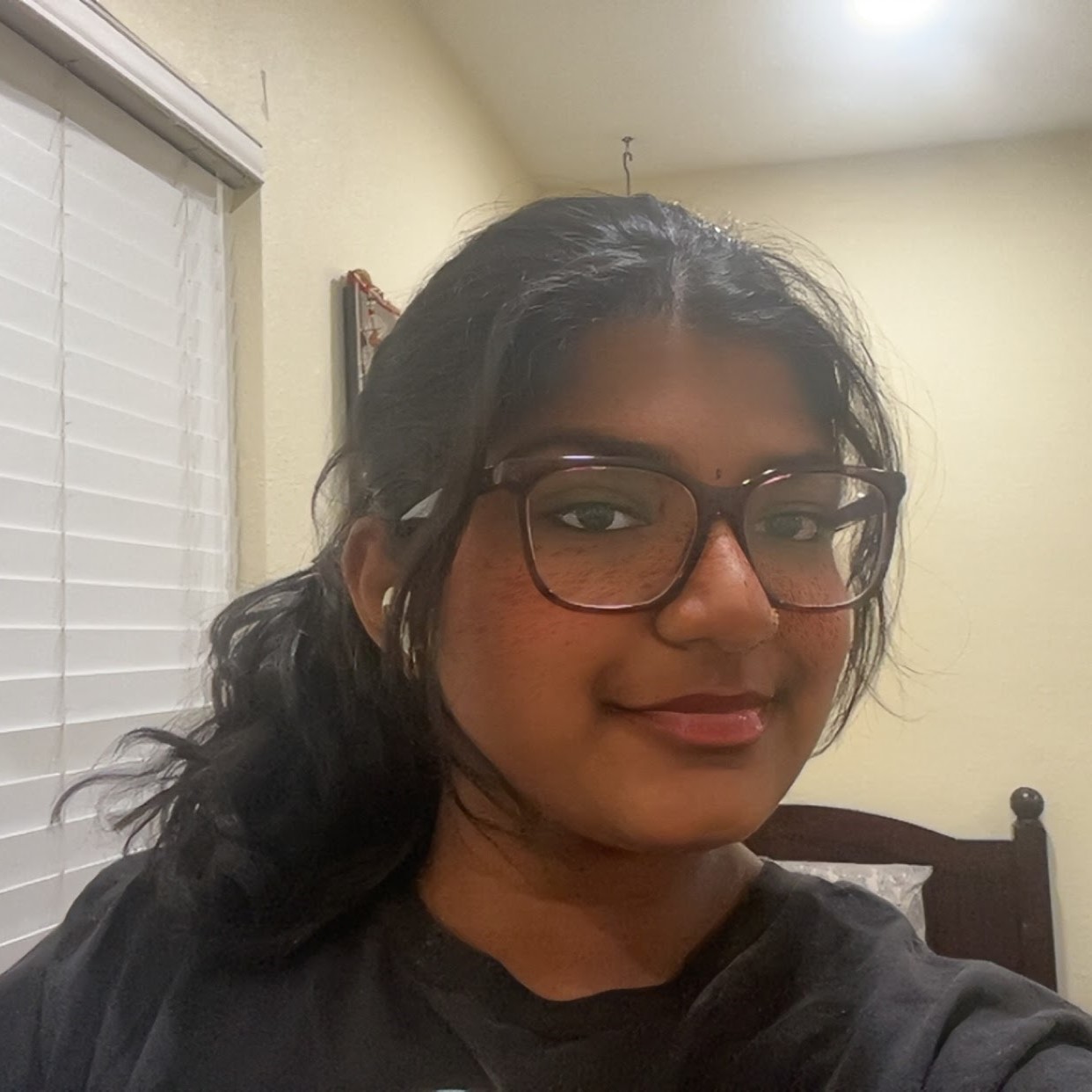 Close-up selfie of Diya wearing large glasses and earbuds, with hair pulled back, indoors near a window with white blinds.