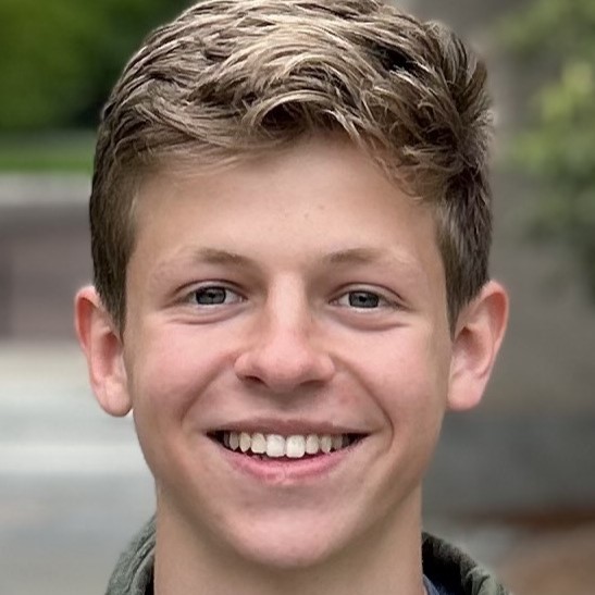 Close-up portrait of Dylan smiling with short light-brown hair against a softly blurred outdoor background.