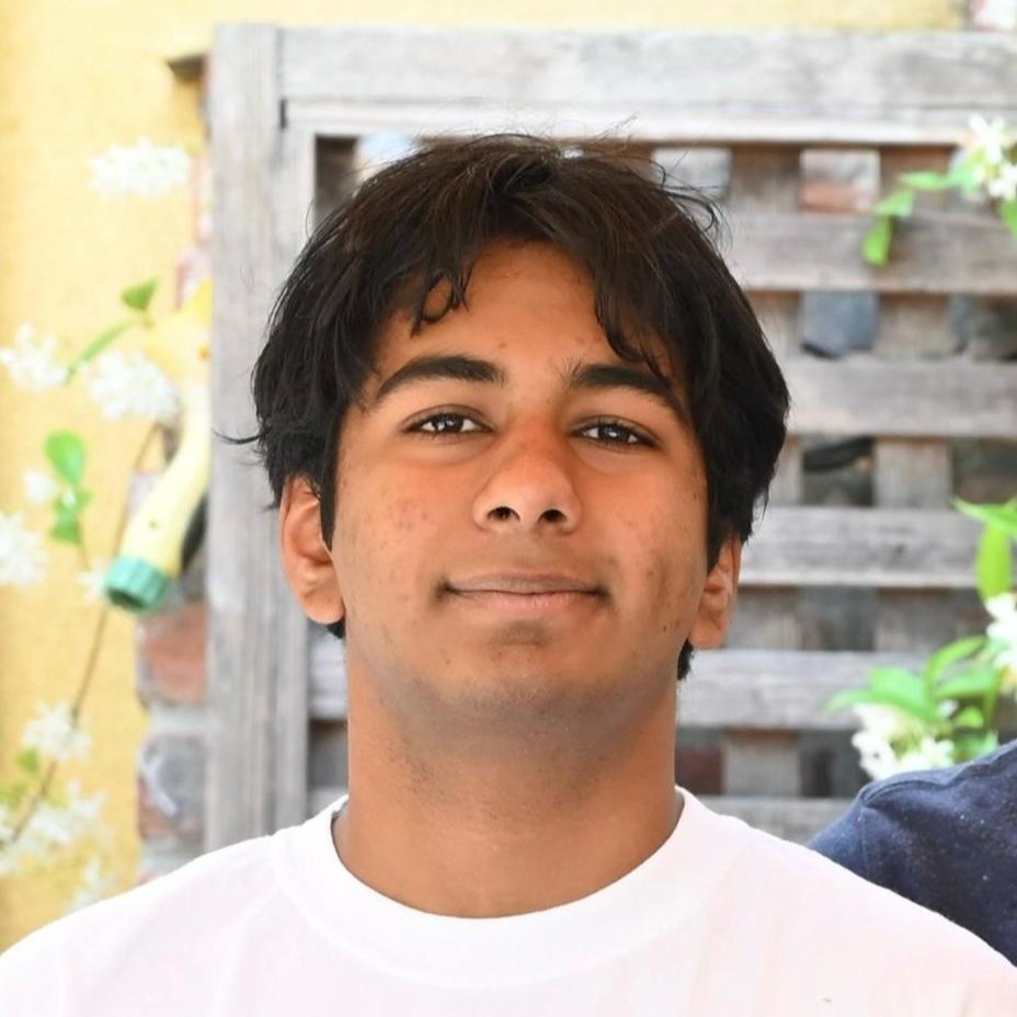 Close-up portrait of Gautam with dark hair wearing a white shirt, standing outdoors in front of a wooden fence and plants.