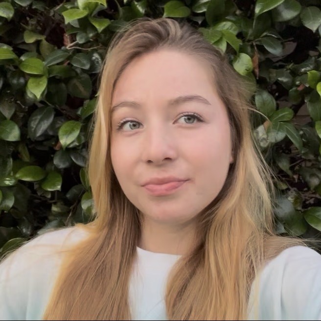 Close-up portrait of Isabella with long blonde hair standing in front of dense green leaves.