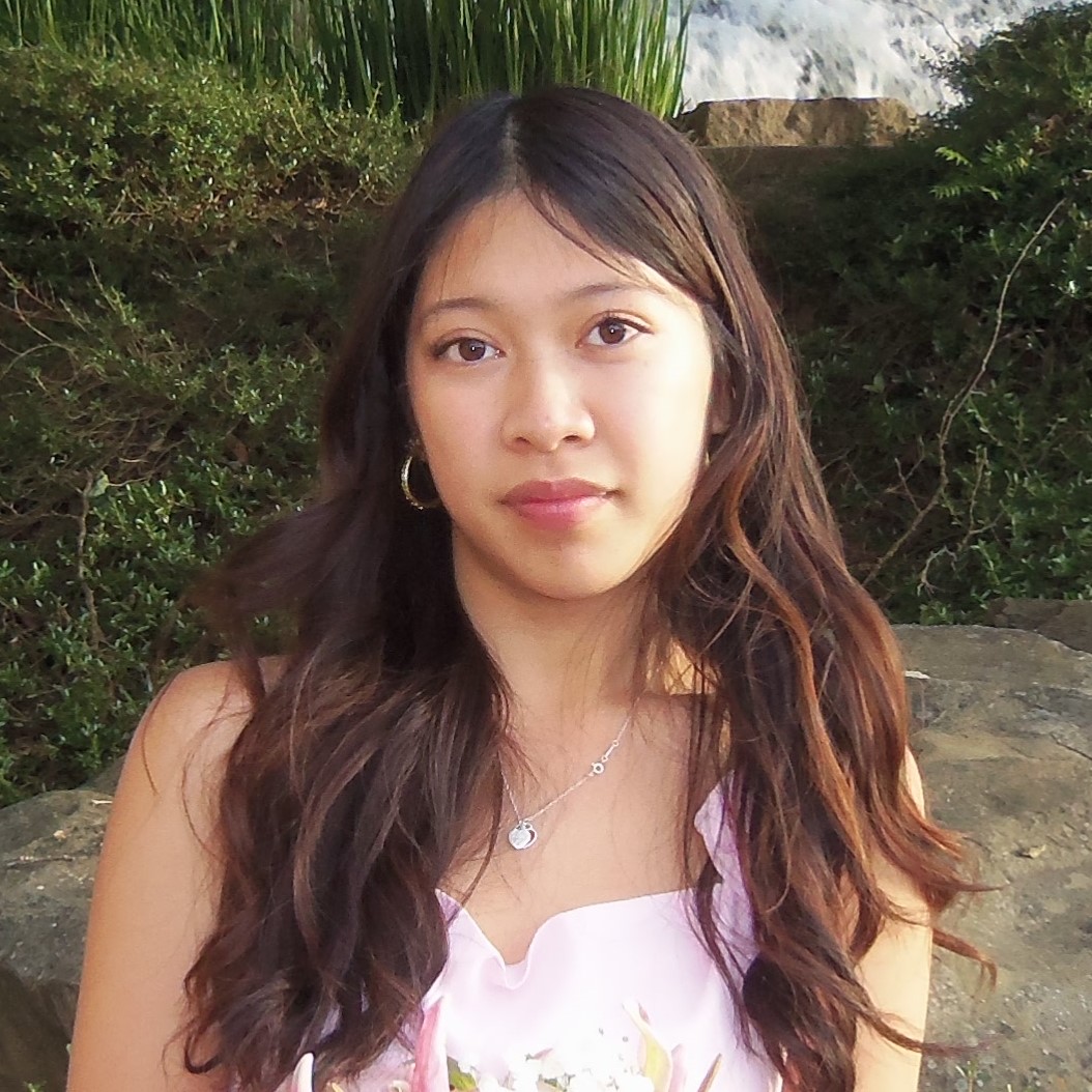 Portrait of Janet with long wavy hair wearing a light pink dress and necklace, standing outdoors in front of greenery and rocks.