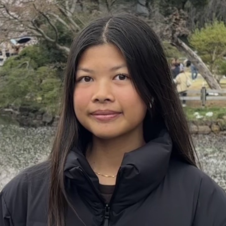 Portrait of Julie with long dark hair wearing a black puffer jacket, standing outdoors with trees and a gravel path in the background.