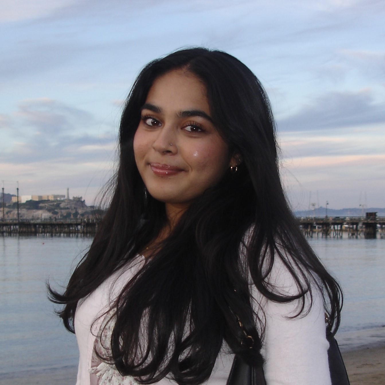 Portrait of Mahiya with long dark hair standing by calm water, with a pier and cloudy sky in the background.