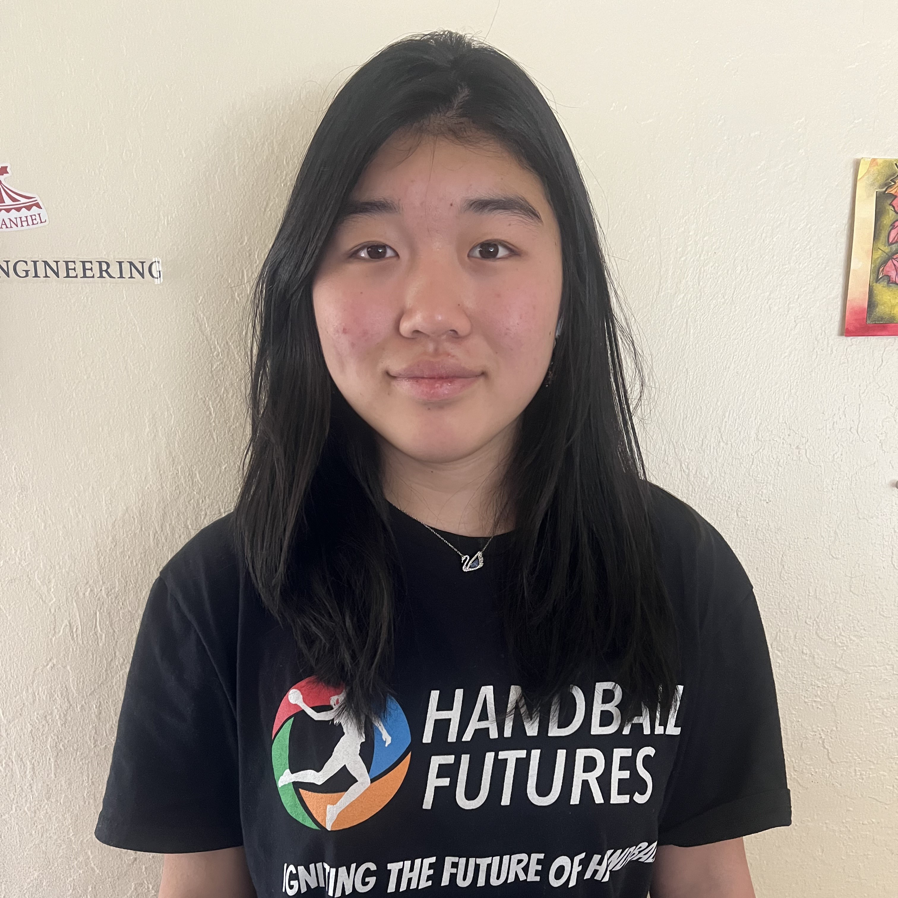 Portrait of a person with long dark hair wearing a black “Handball Futures” T-shirt, standing against a light-colored wall.