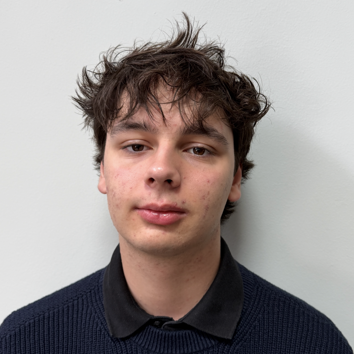 Head-and-shoulders portrait of Marko with tousled dark hair wearing a navy sweater over a black collared shirt against a plain light background.