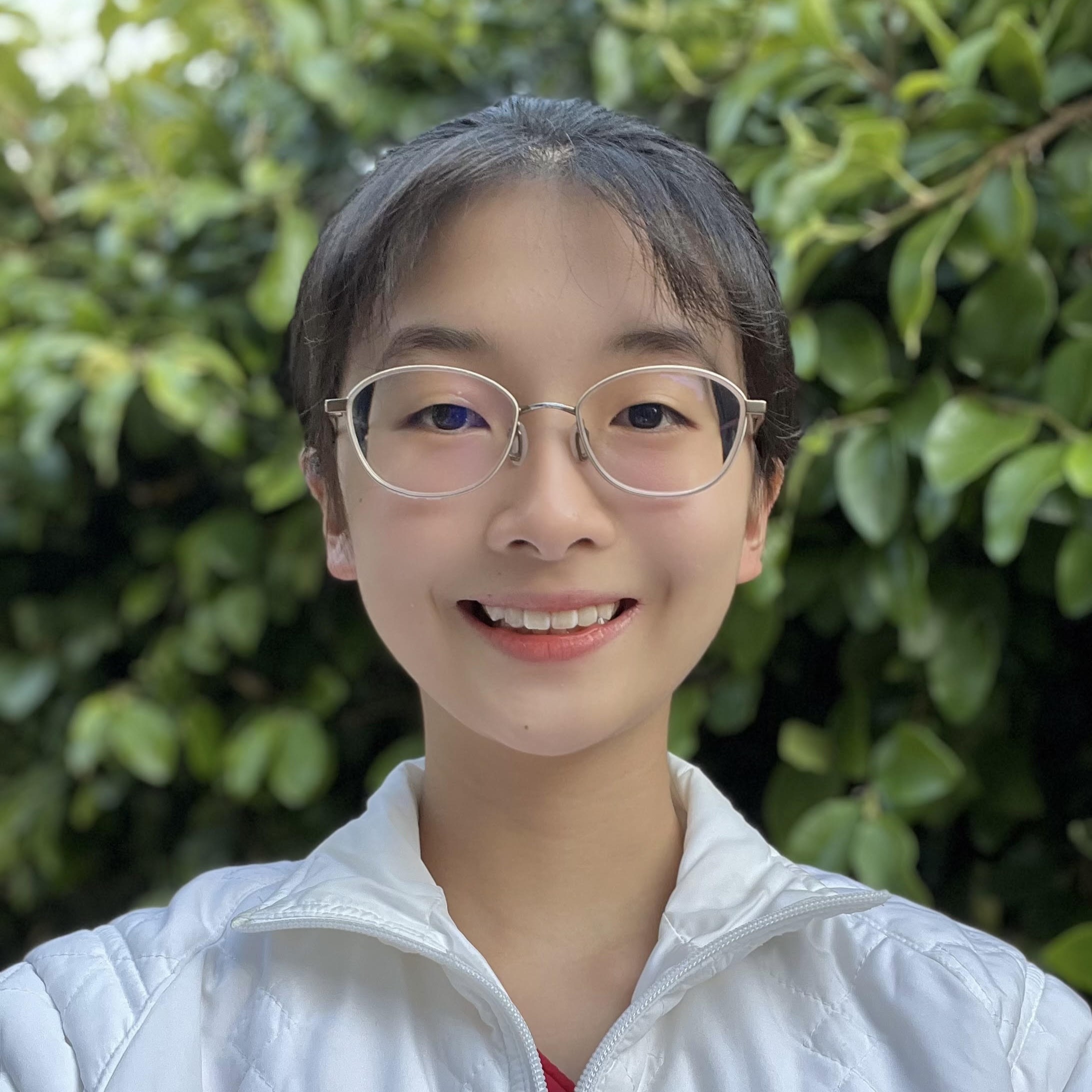 Close-up portrait of Mira wearing round glasses and a white jacket, with leafy greenery in the background.
