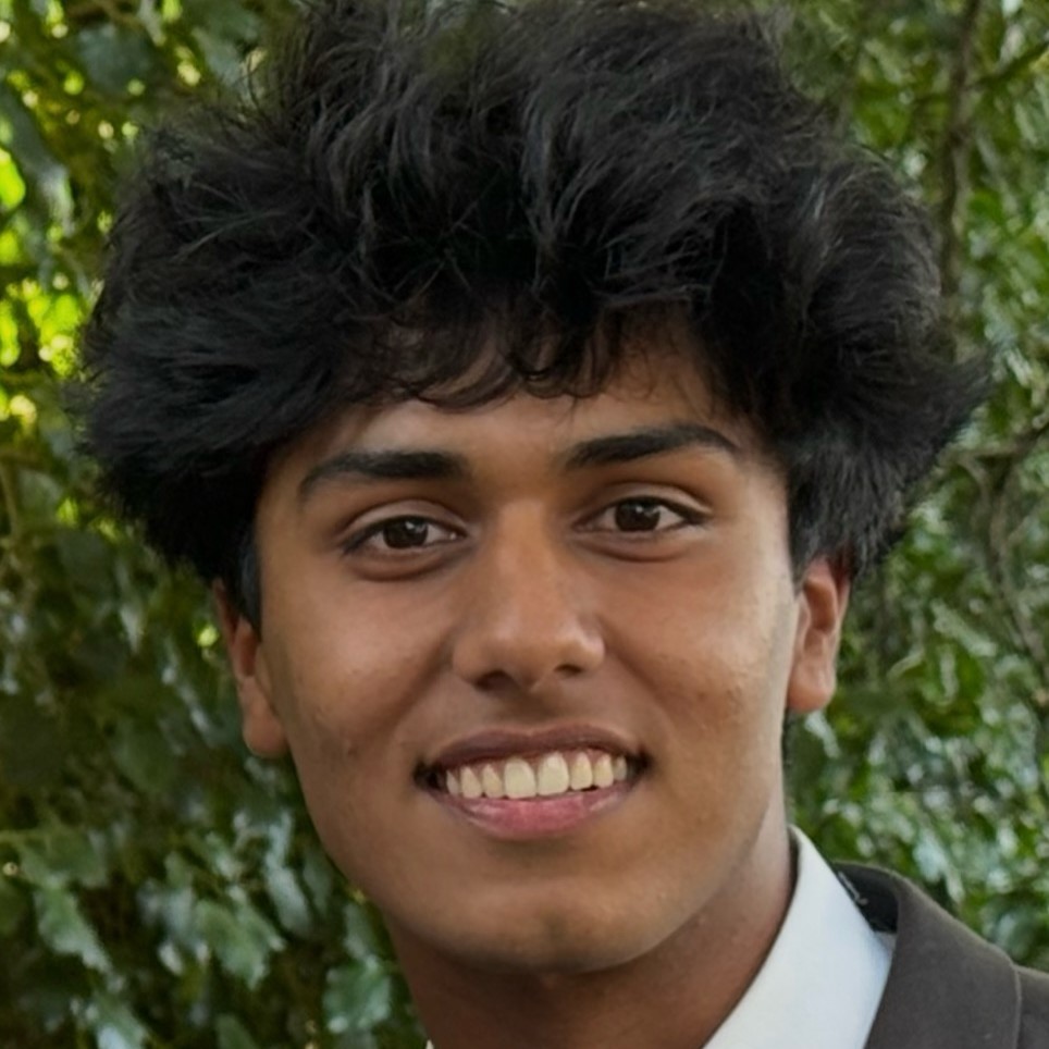 Close-up portrait of Nevaan with thick dark hair, outdoors with green foliage in the background.