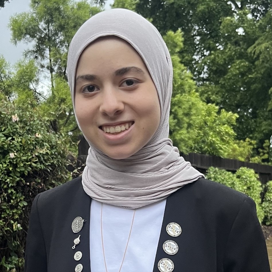 Nour smiling in a light gray headscarf and black blazer with several lapel pins, standing outdoors in front of green trees.