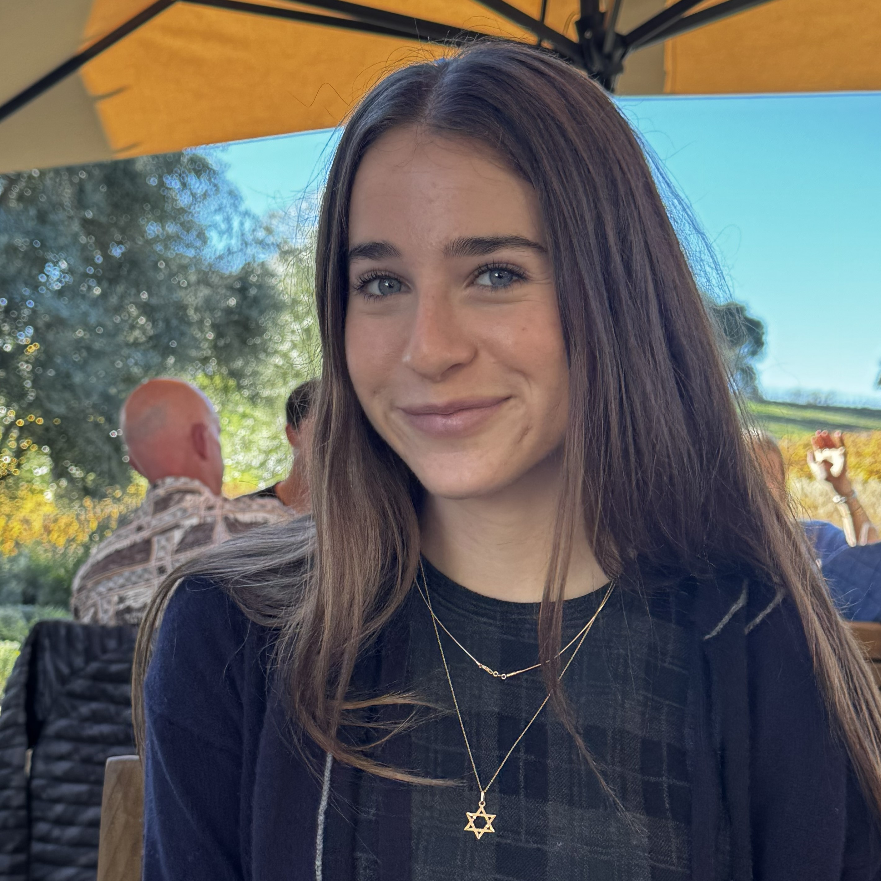 Close-up of Paloma with long brown hair sitting outdoors under a patio umbrella, wearing layered necklaces with a Star of David pendant.