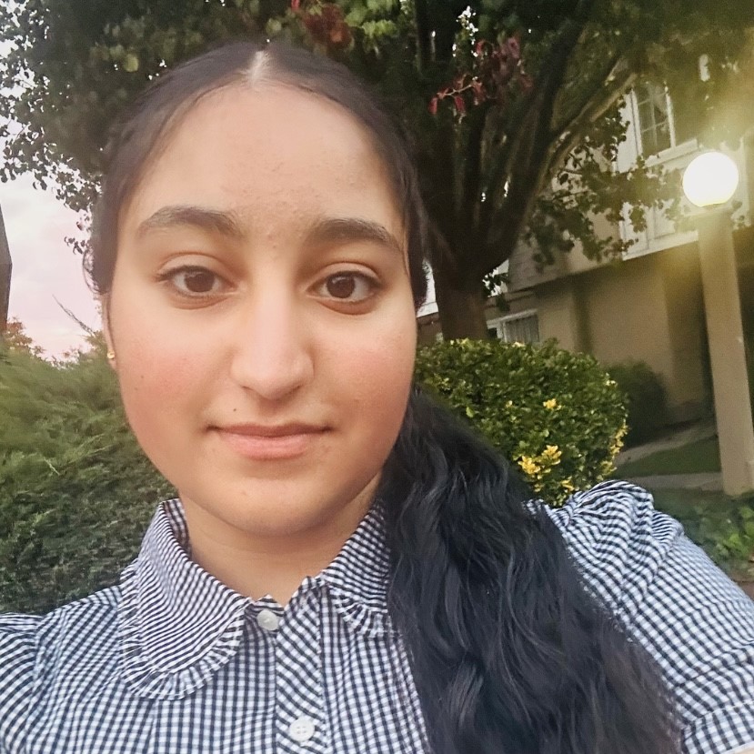 Close-up outdoor selfie of Prabhnoor with long dark hair wearing a black-and-white checkered shirt, with trees, shrubs, and a building in the background.
