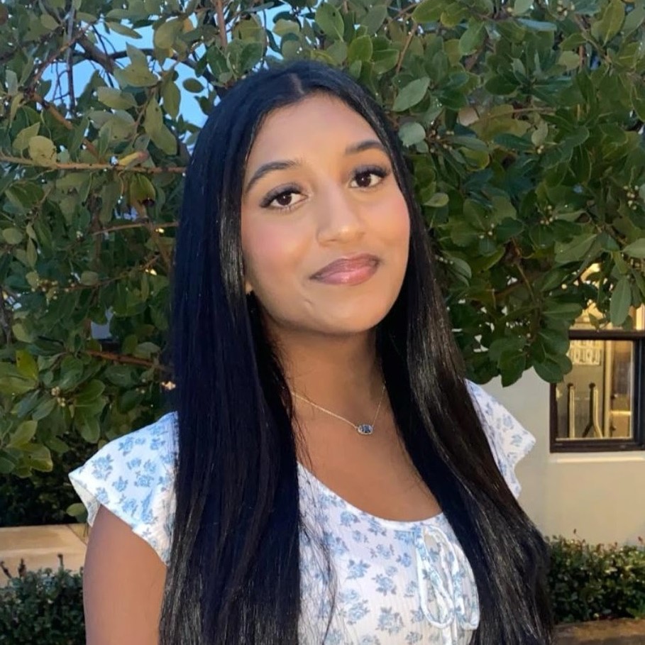 Rothitha with long dark hair wearing a light floral blouse, standing outdoors in front of leafy green trees.