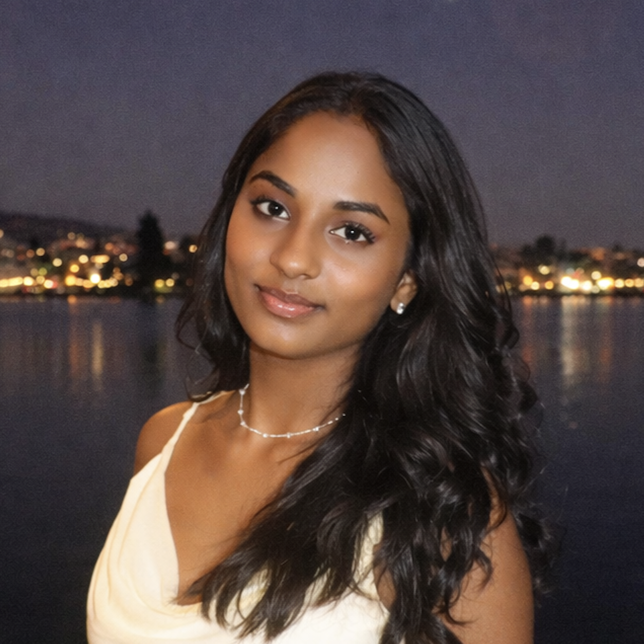 Portrait of Sahaana with long wavy dark hair in a light-colored dress, standing by water at dusk with blurred city lights in the background.
