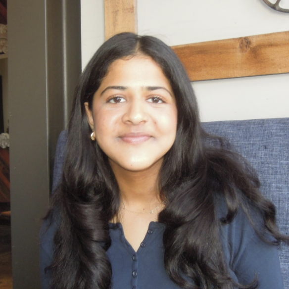 Portrait of Sanchia with long dark hair, wearing a dark top, seated indoors and smiling gently at the camera.
