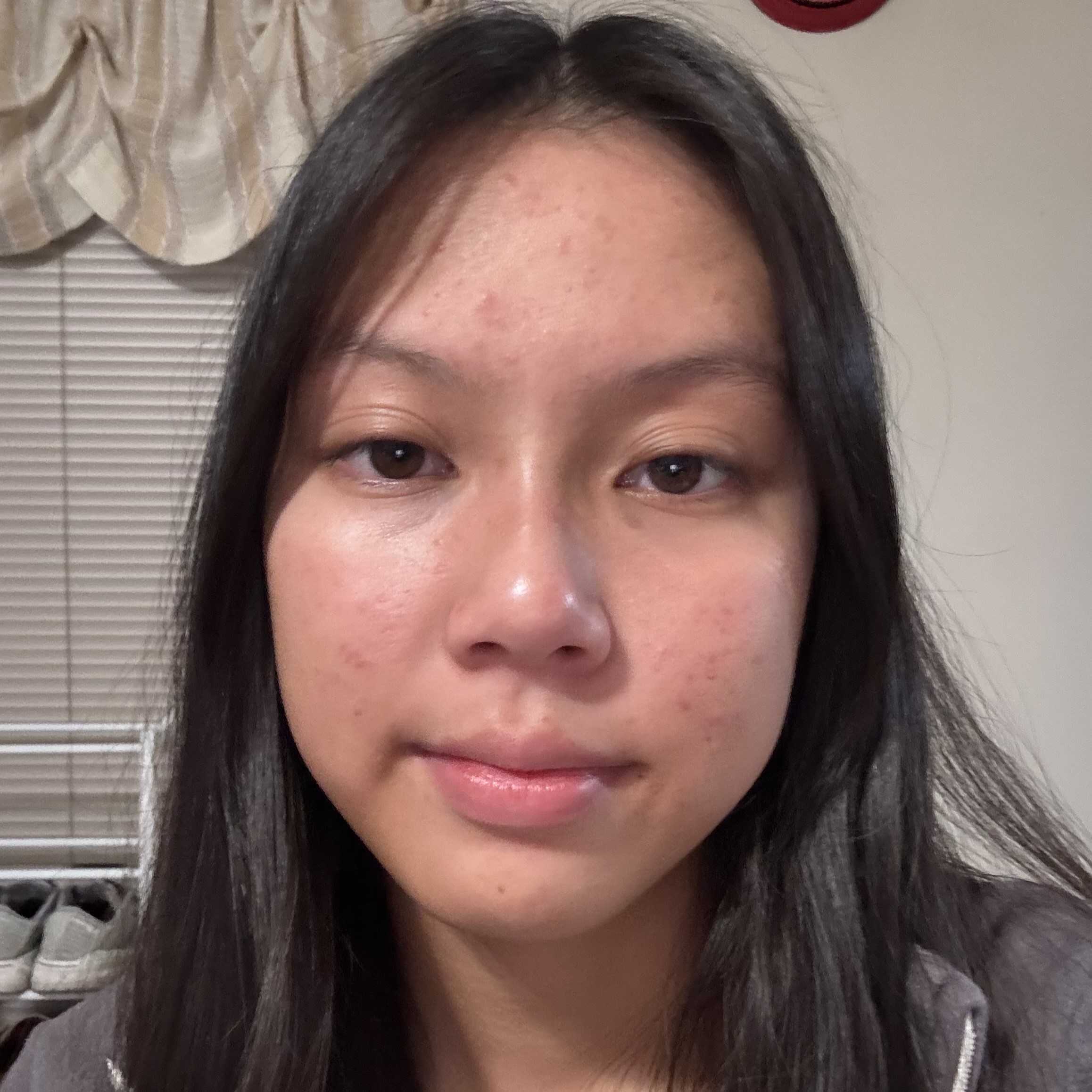 Close-up selfie of Sara with long dark hair, showing a neutral expression indoors with window blinds and a curtain in the background.