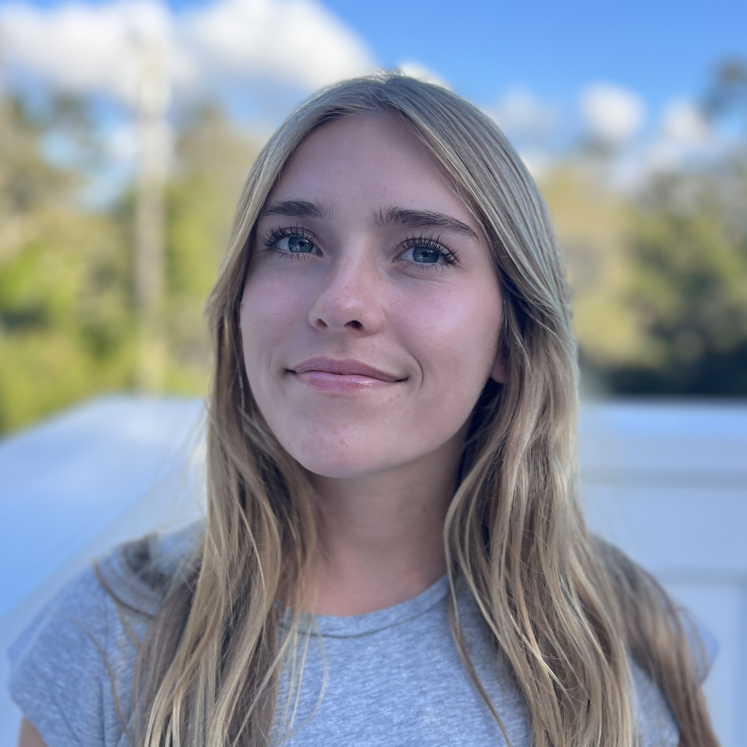 Close-up portrait of Tatum with long blonde hair outdoors, with a blurred lake, trees, and blue sky in the background.