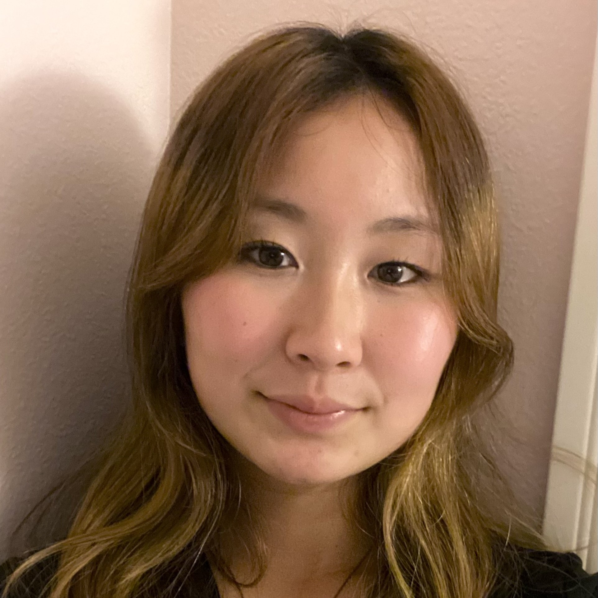 Close-up selfie of Tina long light-brown hair and subtle makeup, looking at the camera against a plain indoor wall.
