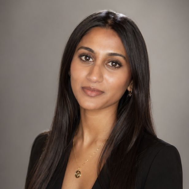 Studio headshot of Varya with long dark hair wearing a black blazer and a gold necklace against a gray background.