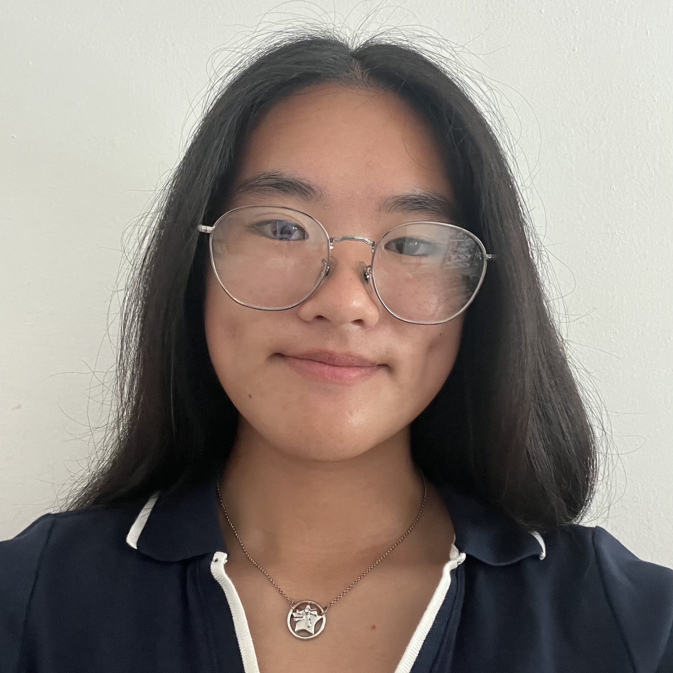 Close-up portrait of Vivian with long dark hair wearing round glasses and a necklace, smiling slightly against a plain light wall.