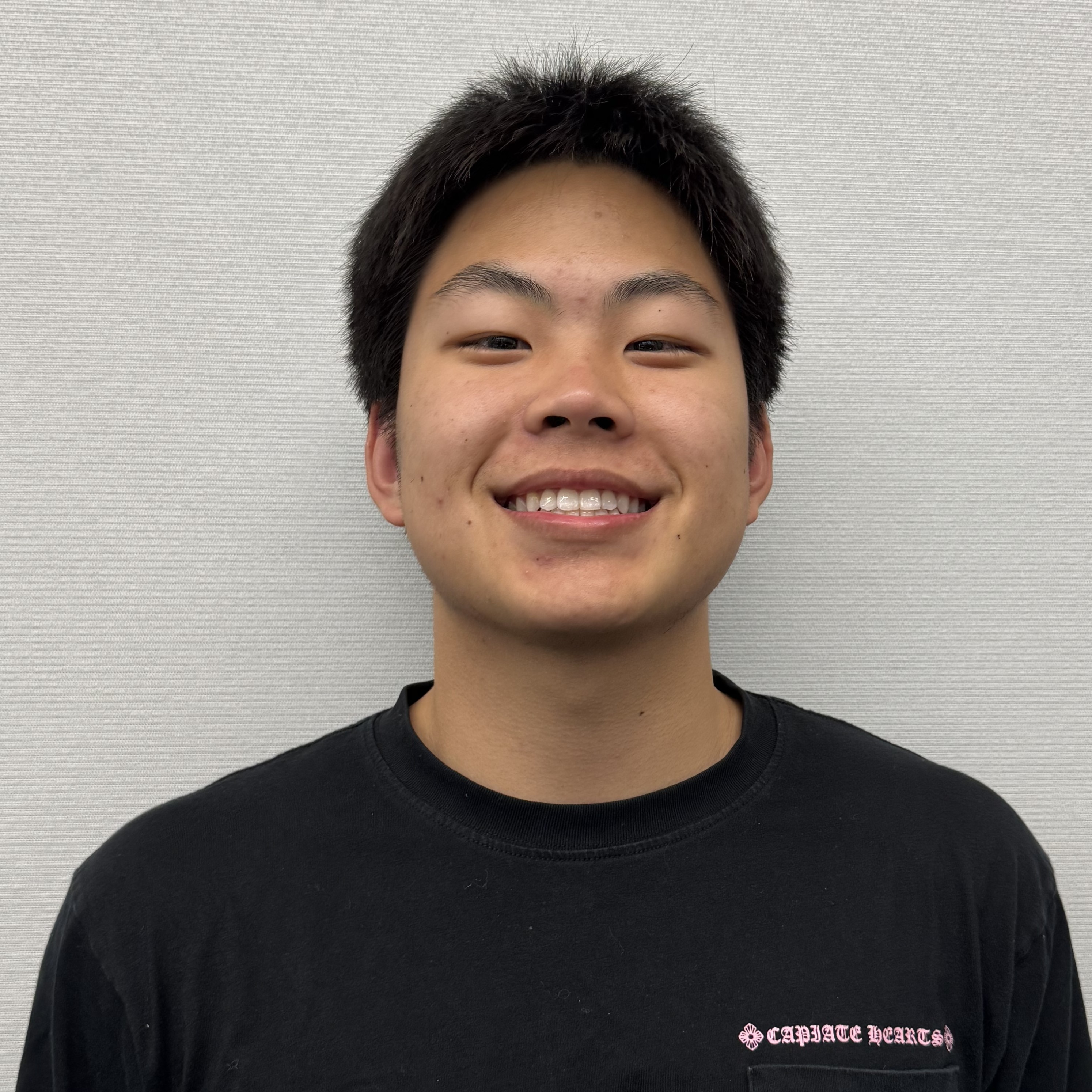Close-up portrait of a smiling person wearing a black shirt against a light gray textured wall.