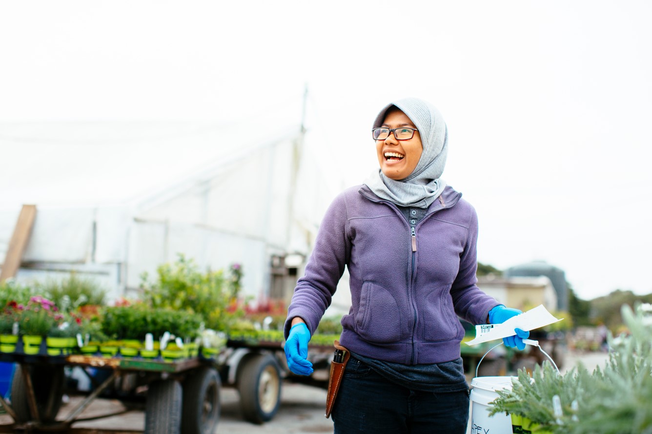 Image of a student smiling and wearing a hijab, glasses, and blue gloves and standing in a garden.