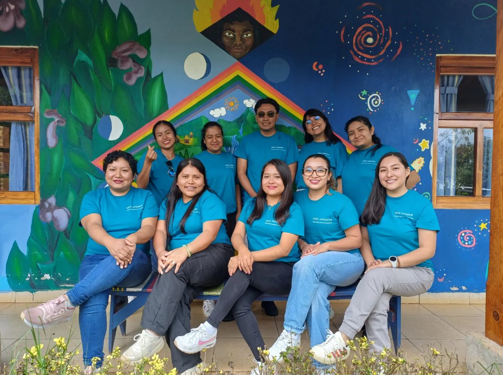 Group photo of Mexico Leadership Program cohort in matching teal HEAL t-shirts posing on a bench in front of a colorful mural with a rainbow and stars.